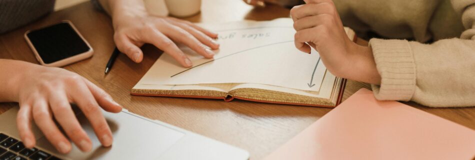 Two people collaborating on a sales growth strategy, one using a laptop and the other pointing at a graph in an open notebook.