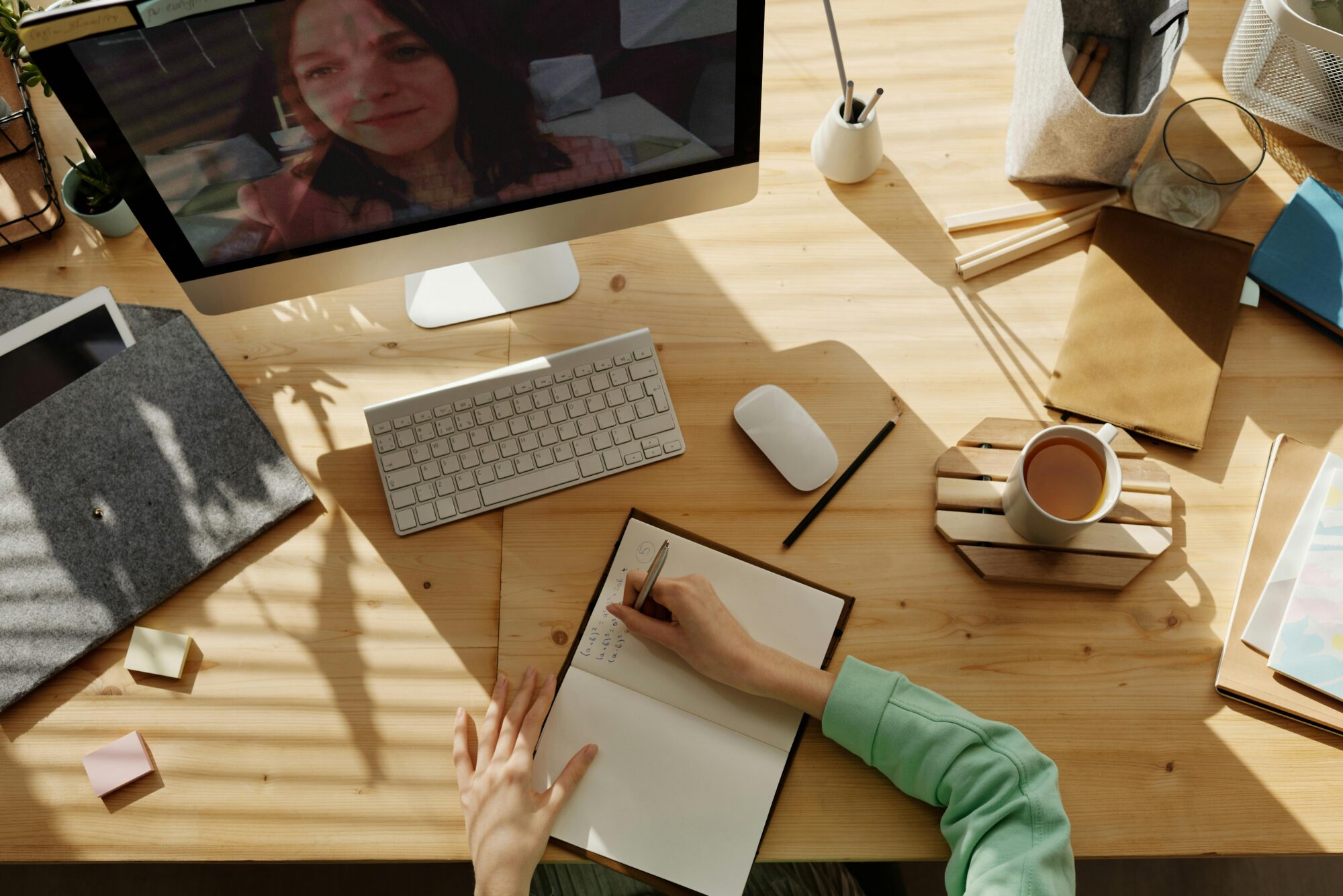 A workspace with a person taking notes during a virtual meeting, with a computer, notepad, and coffee on the desk.