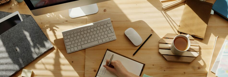 A workspace with a person taking notes during a virtual meeting, with a computer, notepad, and coffee on the desk.