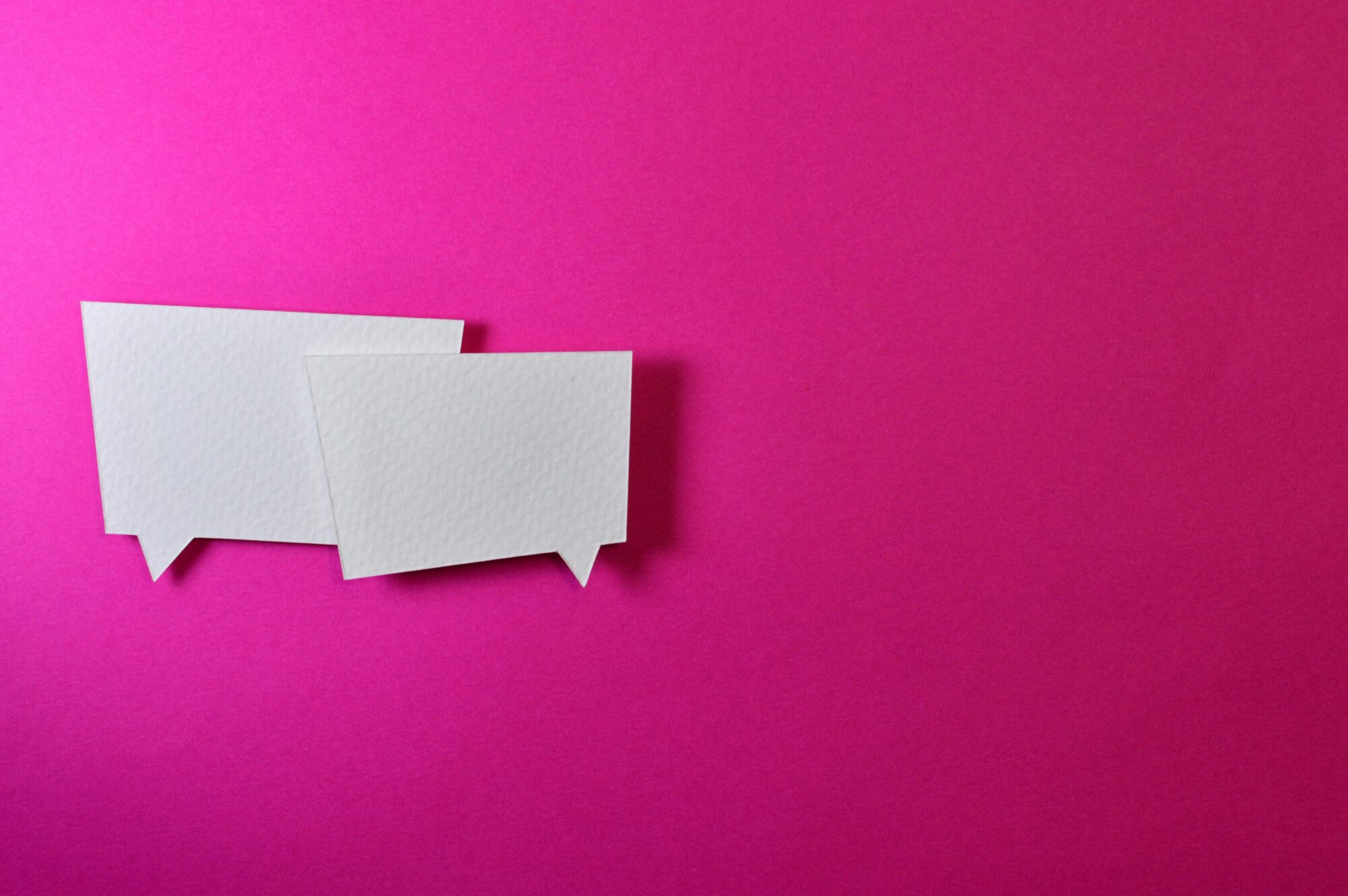 Two white paper cutouts shaped like chat bubbles placed on a bright pink background, symbolizing communication or conversation.