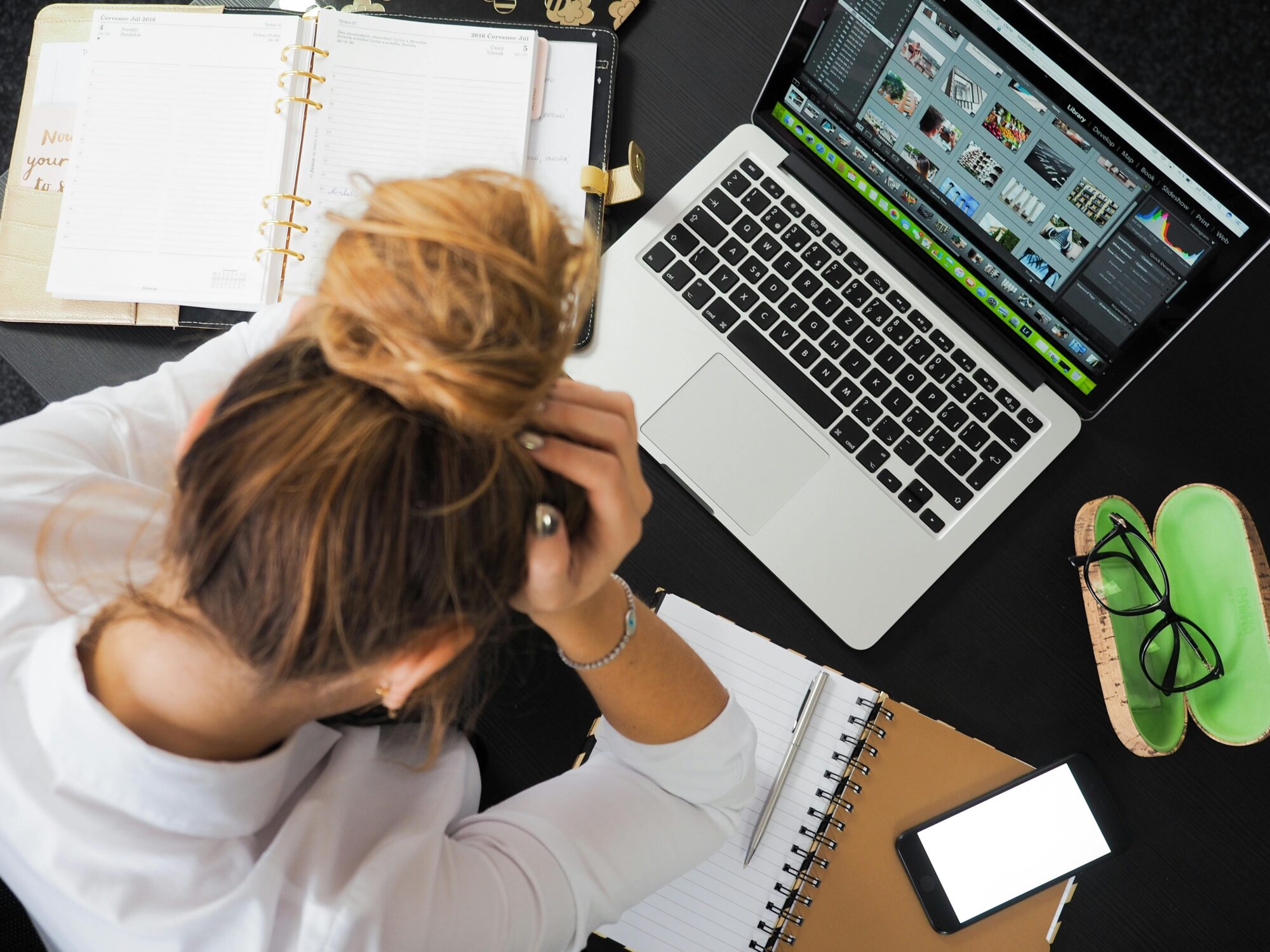 A woman sitting at her desk holding her head, surrounded by notebooks, a laptop, and a phone, appearing stressed.